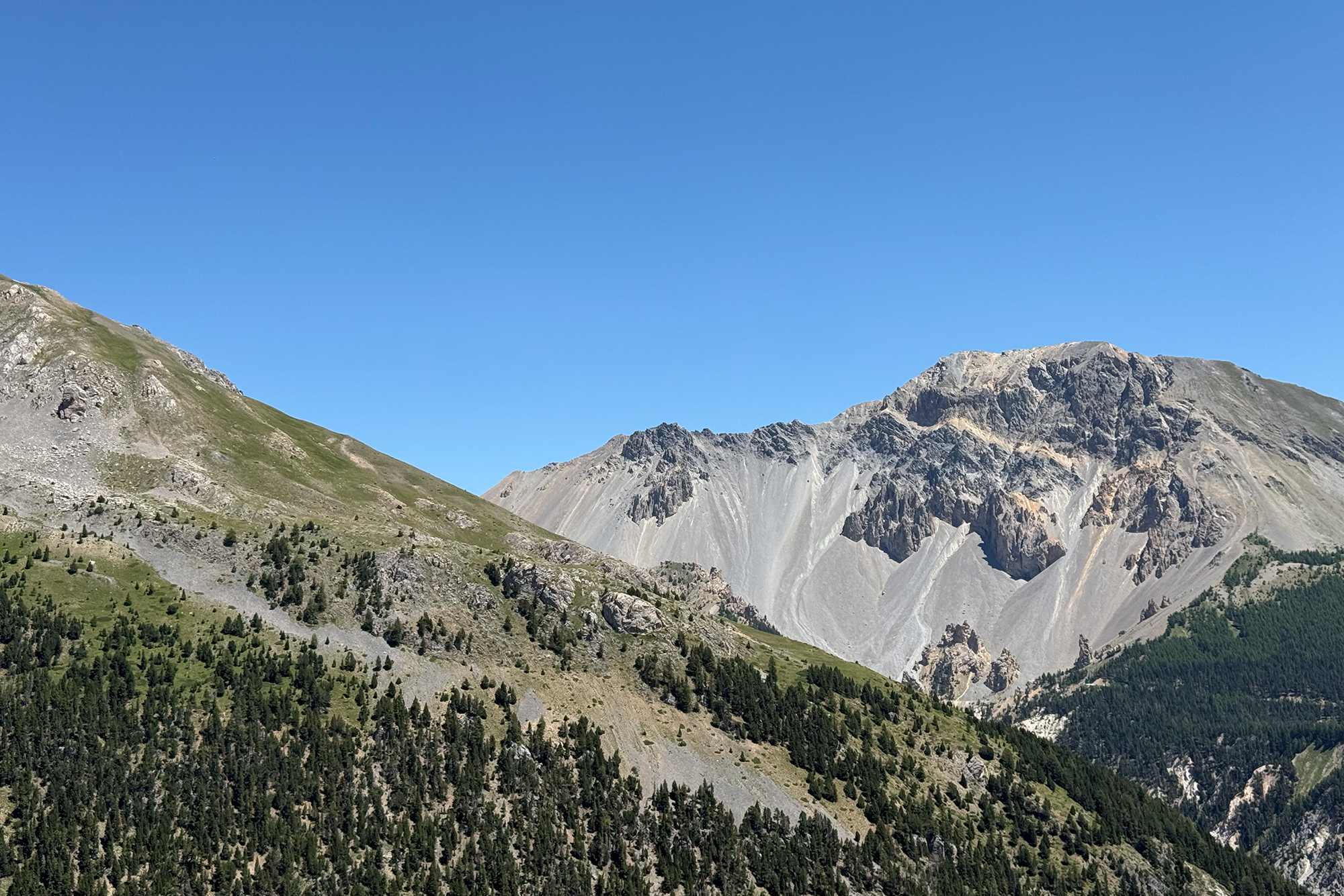 Breathtaking mountain landscape with rugged gray peaks under a clear blue sky and green forested lower slopes in the foreground.