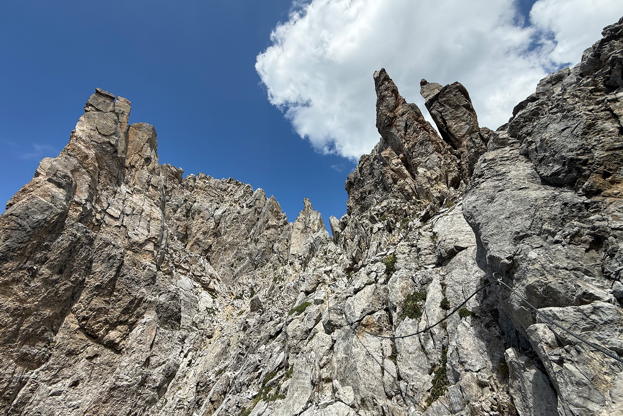 Jagged rocky cliffs rise against a blue sky with white clouds; a safety rope runs along the slope below.