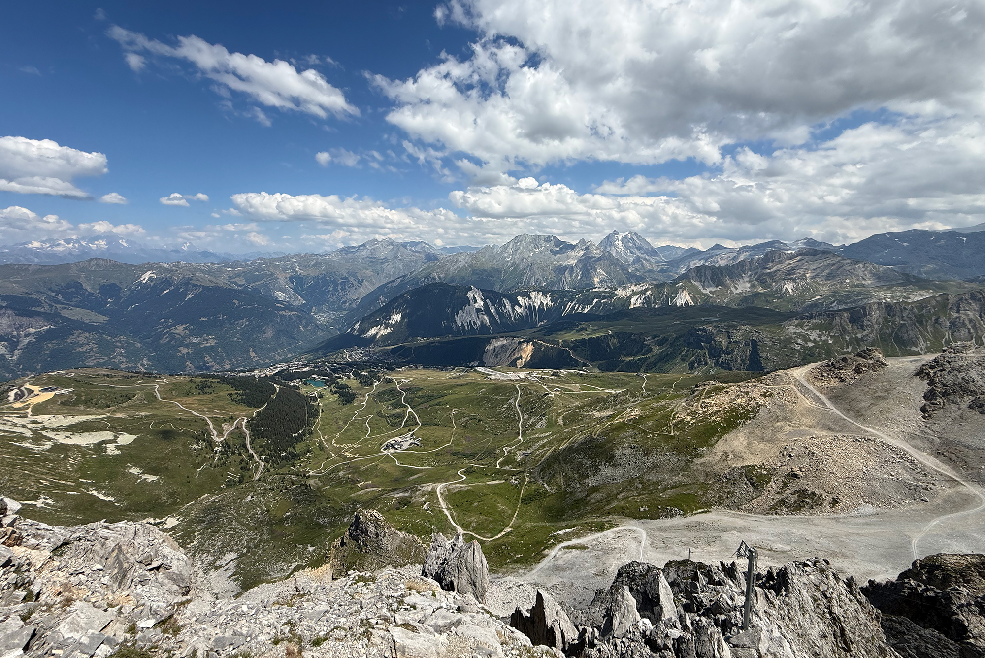 Mountain valley with winding trails, rocky foreground and distant snow-capped peaks under a partly cloudy sky