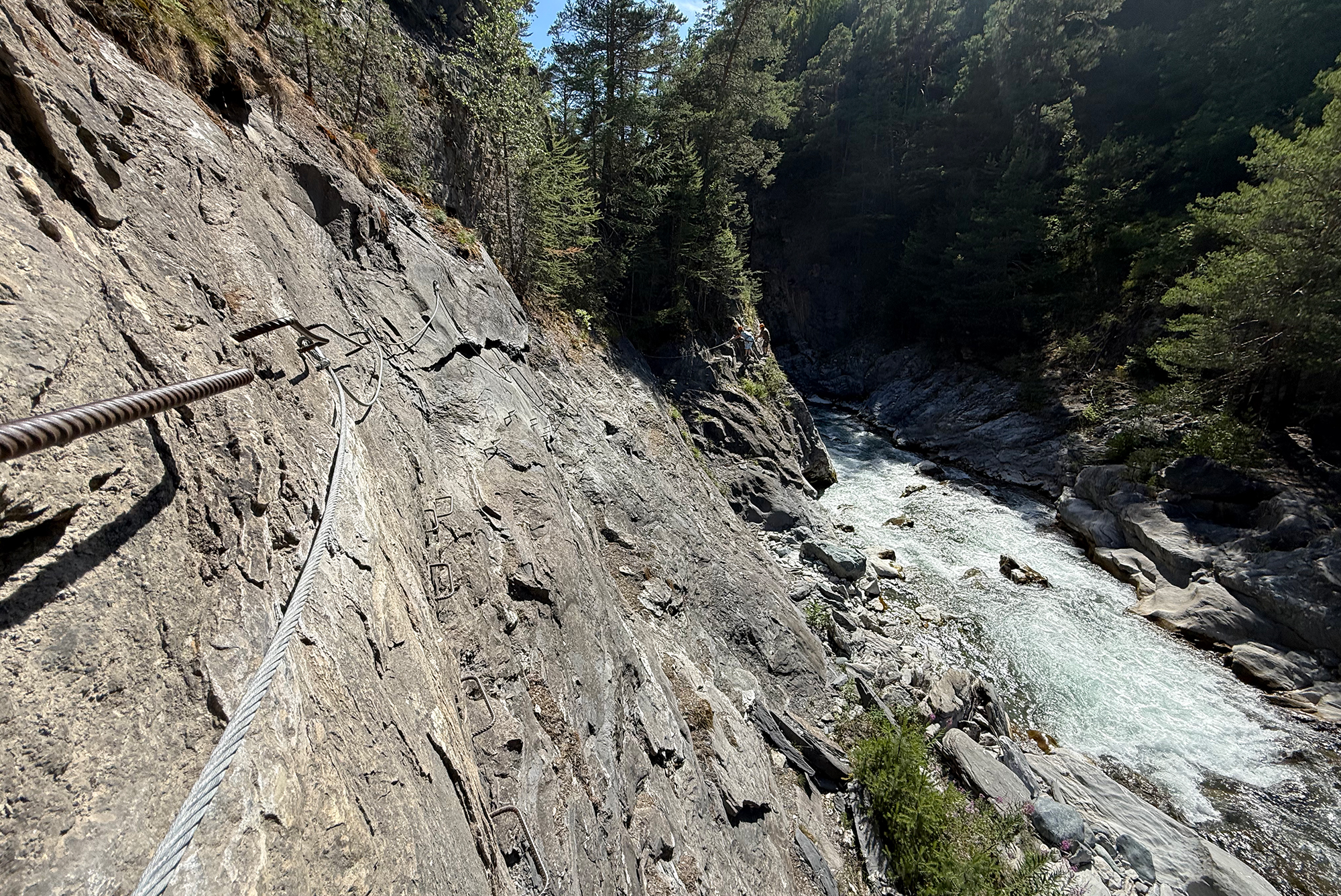 Steep rock cliff with a metal fixed ladder and steel cable, a river raging below and dense forest on both sides; hikers in the distance.