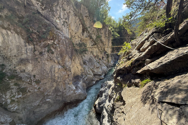 Narrow rocky canyon with a rushing river and hikers crossing a rope-and-plank bridge between cliffs.