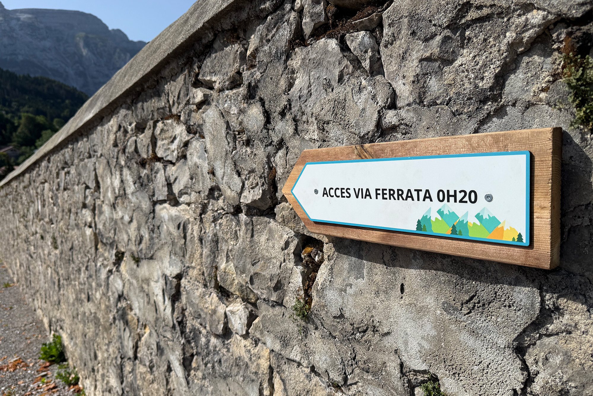 Wooden sign attached to a rough stone wall reading 'ACCES VIA FERRATA OH20' with mountain graphics at the bottom.