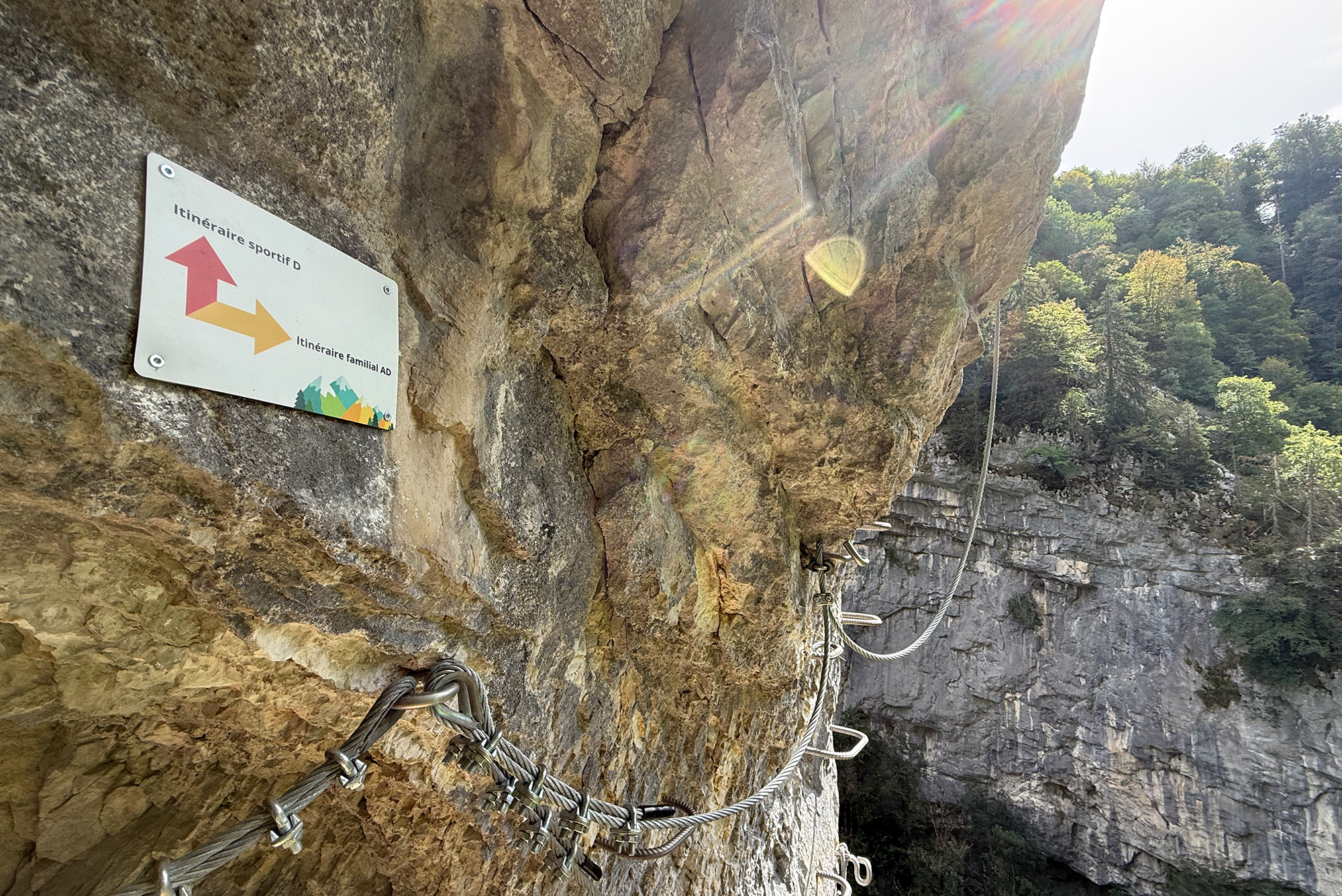 Rock cliff with a via ferrata setup: metal cables, anchors, and carabiners along the rock face, with a forested canyon below.
