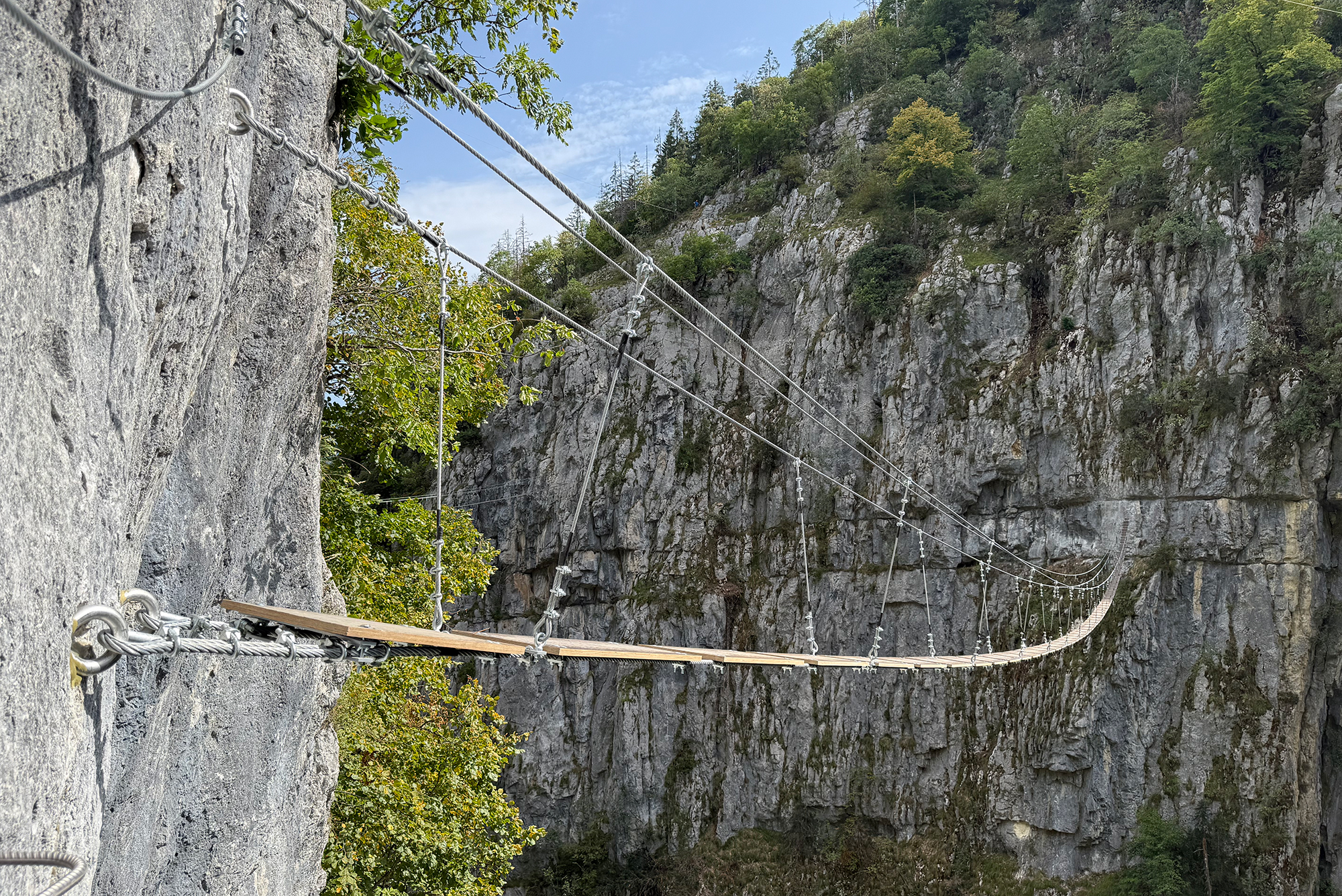 Suspension rope bridge spanning a rocky gorge, anchored to a cliff with metal cables and trees in the background.
