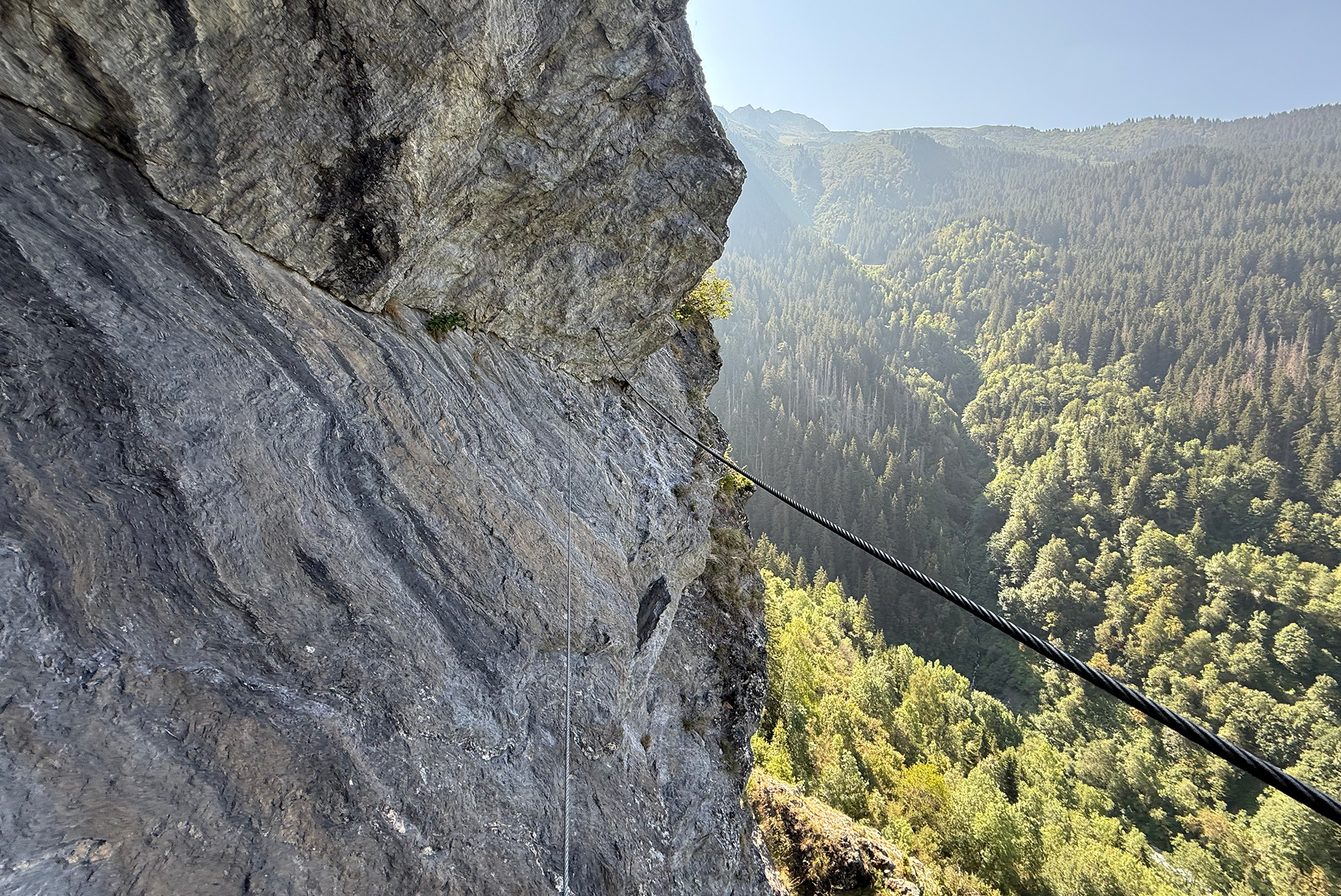 Steep rock cliff with a safety cable descending along the rock face toward a forested valley below.