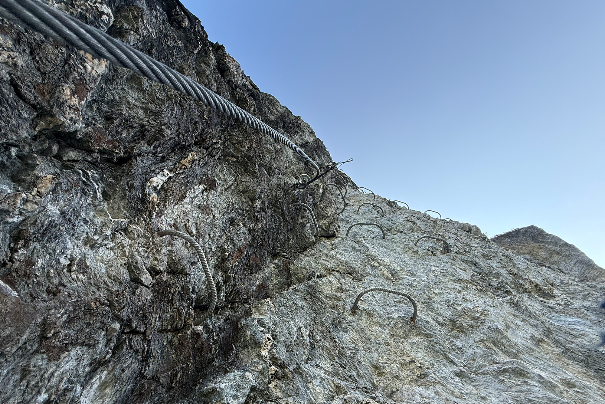 Rocky cliff face with attached steel cables and hooks for climbing against a clear blue sky