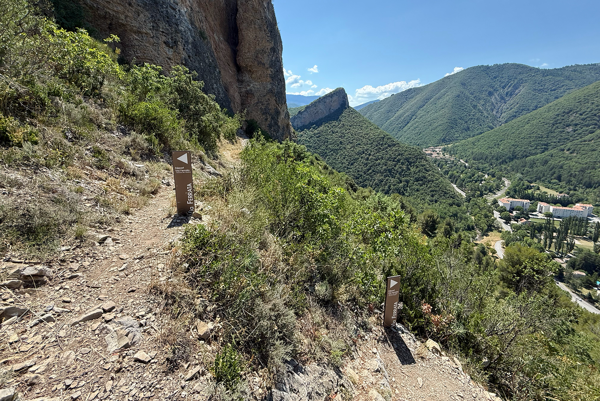 Steep rocky hiking path along a cliff with green shrubs and a Via Ferrata sign, overlooking a valley and village below.