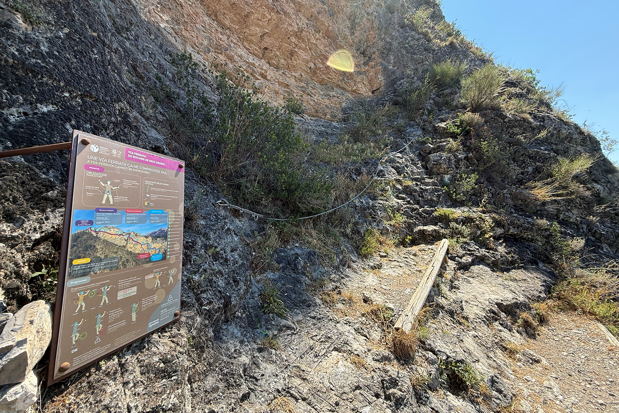 Informational signboard about a via ferrata mounted on a rocky cliff, with a map and safety instructions along a mountain trail.