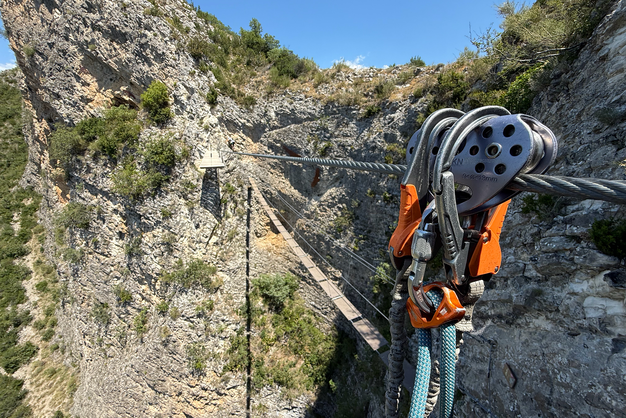 Climber's view: metal pulley and blue rope attached to a steel cable over a rocky canyon, with a suspended wooden bridge below and rugged cliffs nearby.