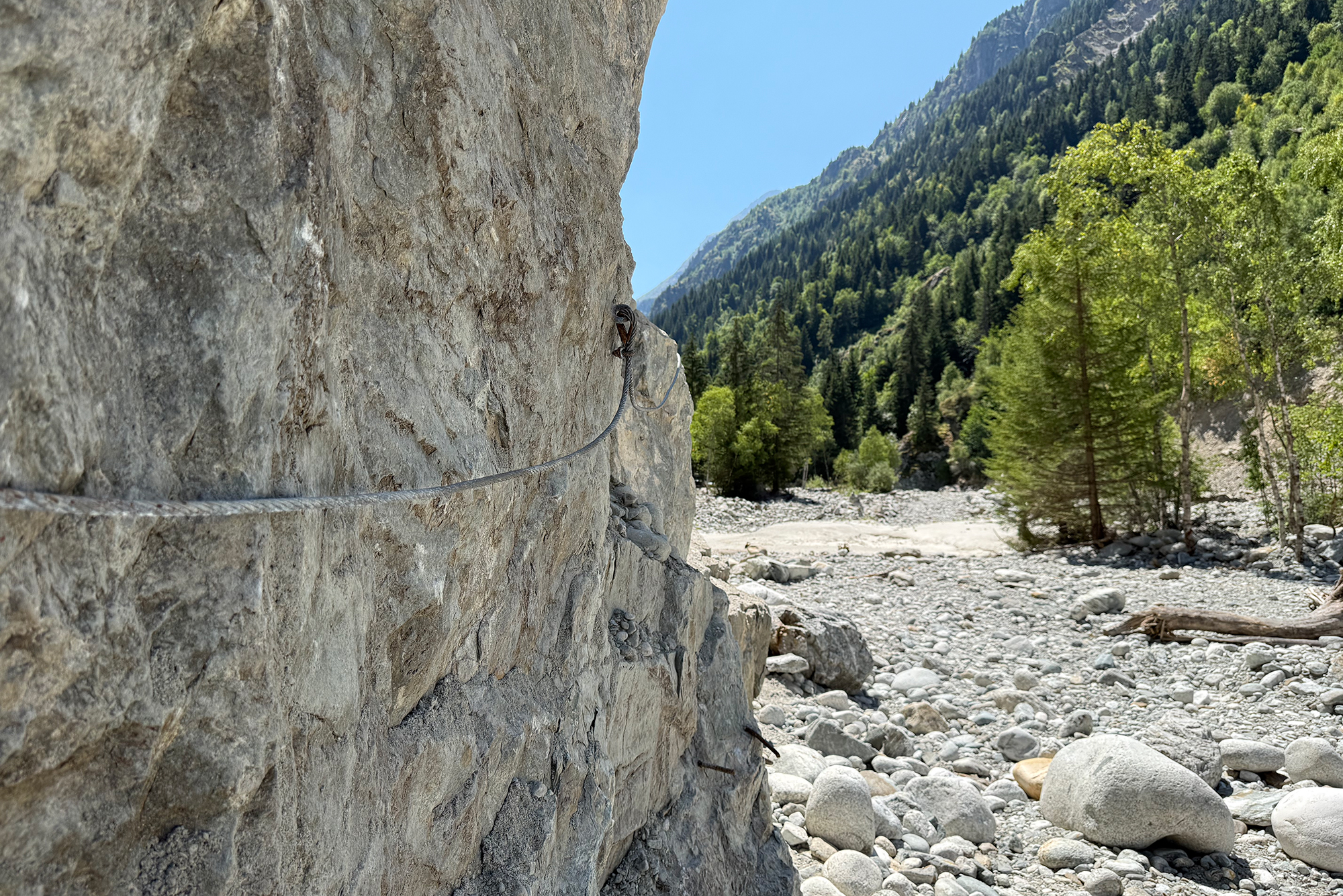Rock wall with an anchored rope for climbing, beside a rocky riverbed and forested mountains in the distance.