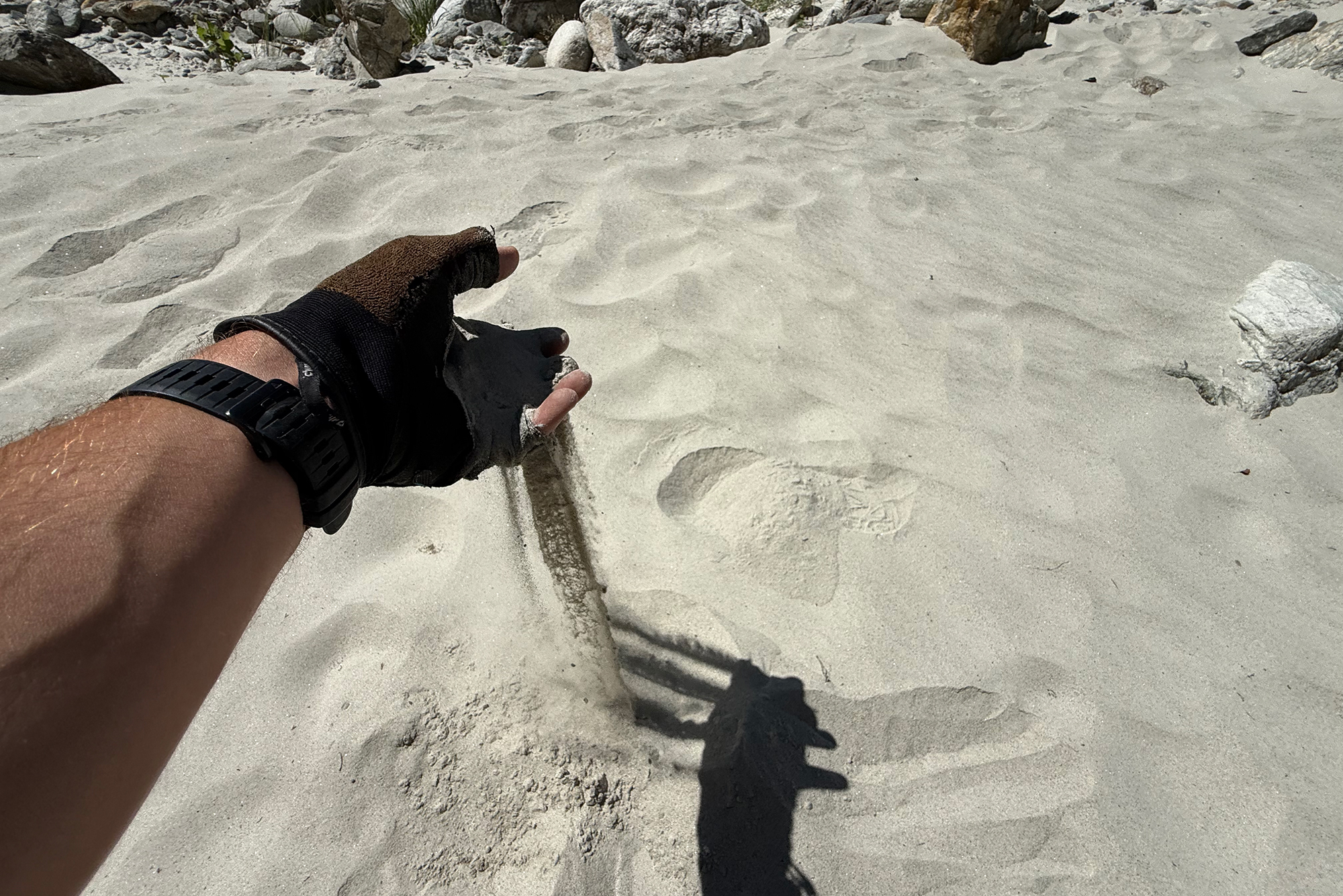Gloved hand digging a narrow trench in light sand at a rocky beach, with a wristwatch visible on the wrist.