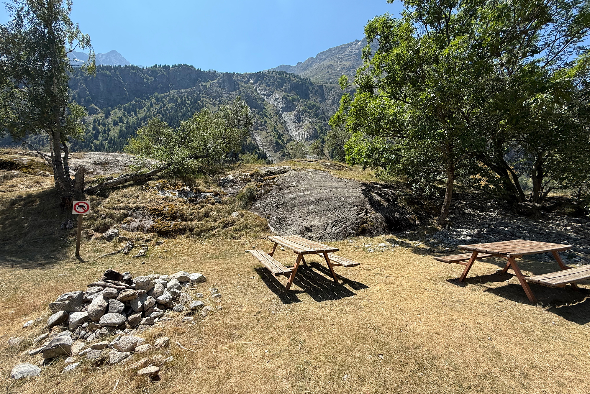 Picnic area in a rocky, sunlit meadow with two wooden tables, trees on both sides, and a mountain range in the background; a small no-dogs sign is visible on a post to the left.
