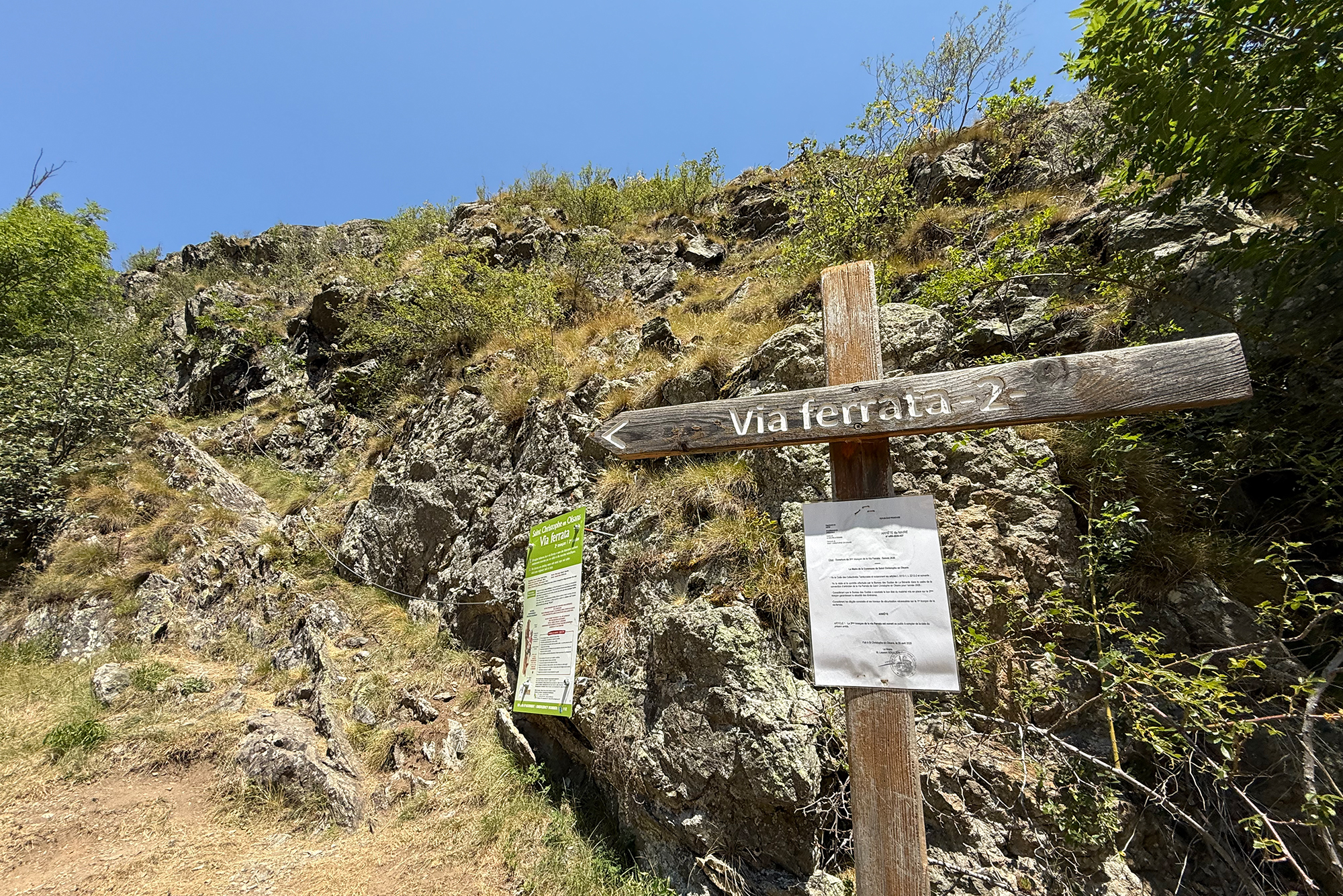 Wooden signpost reading 'Via ferrata 2' at a rocky hillside with trail notices below under a blue sky.