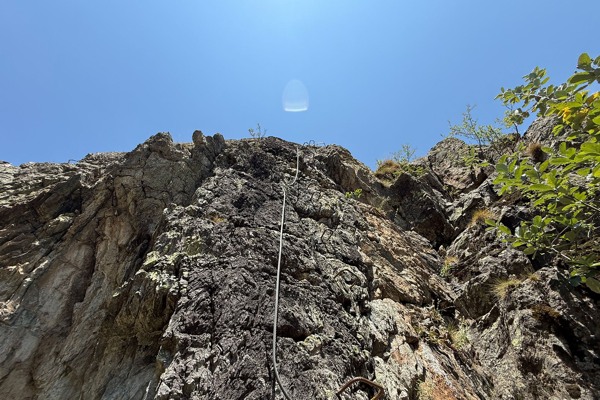 Jagged rock cliff with a climbing rope running down the center under a bright blue sky; vegetation at sides.
