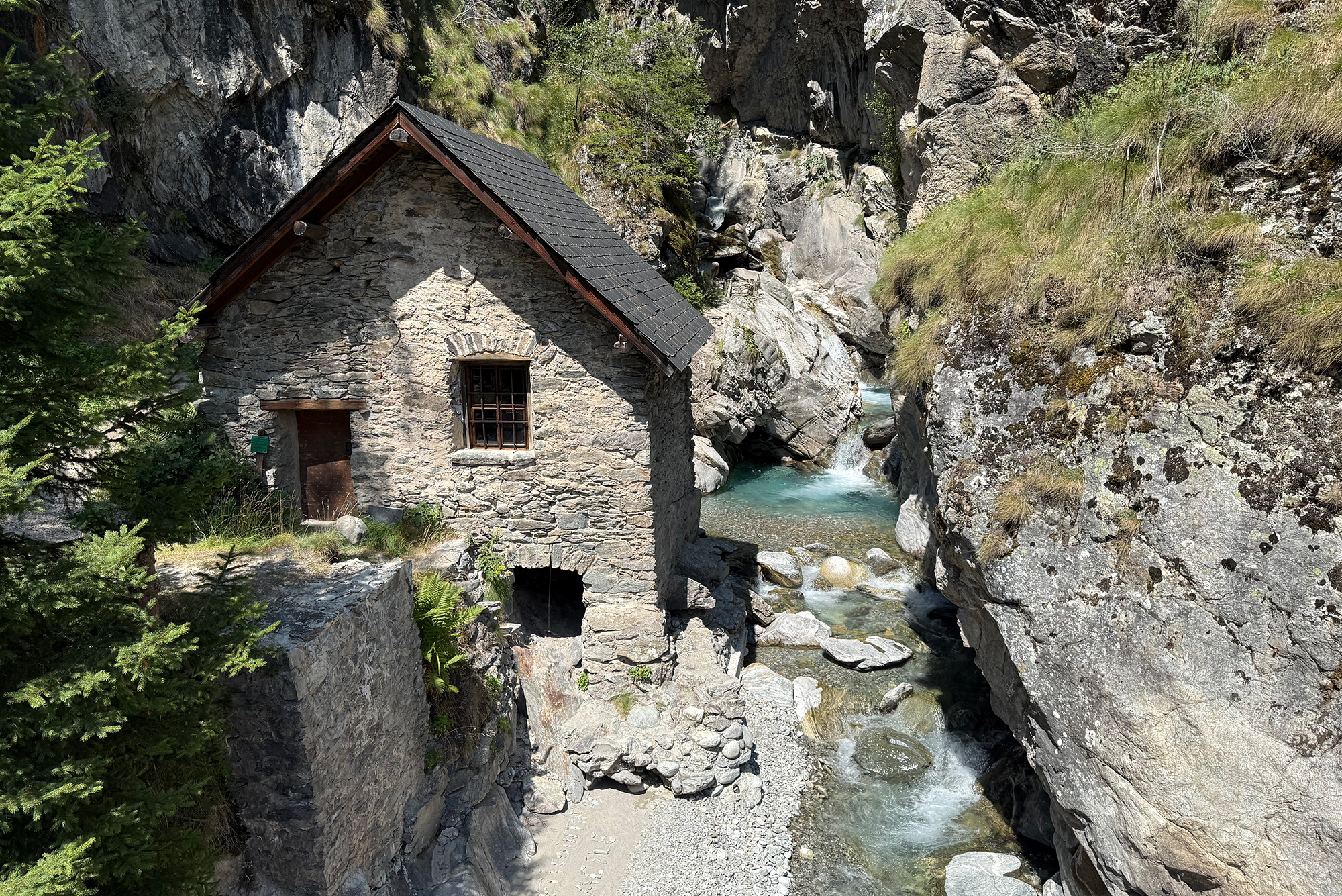 Stone cottage with a slate roof built into a rocky canyon beside a turquoise river, surrounded by cliffs and greenery.