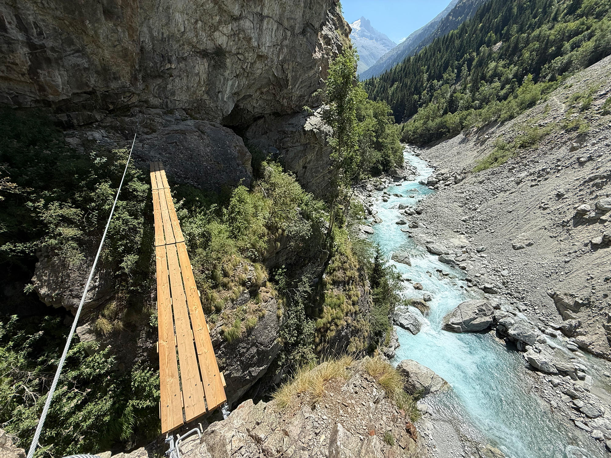 Wooden suspension footbridge spanning a rocky gorge with a turquoise river below and steep cliffs on both sides, mountains in the distance.