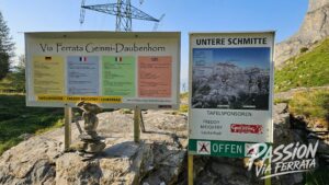 Alpine information boards showing Via Ferrata Gemmi-Daubenhorn and Untere Schmitte signs with a map and sponsor logos, mounted on metal stands on rocky ground.