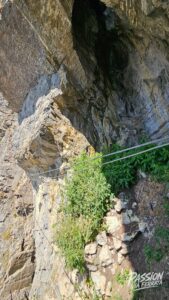 Rugged cliff face with gray-brown rock and green plants growing from cracks; metal safety cables run along the path beside the rock wall.