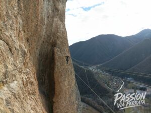 Climber wearing a helmet on a rock face, secured to a metal ladder and cables along a cliff with mountain valley below.