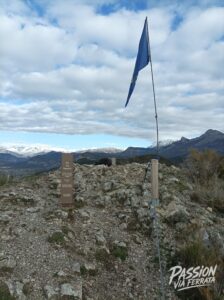 Flagpole with a blue flag on rocky terrain, mountains in the background under a cloudy sky; a trail sign nearby.