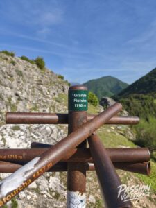 Green sign reading 'Grande Fistoire 1110 m' mounted on a rusted metal barrier, with rocky hillside and distant mountains under a blue sky.