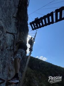 Climber wearing a helmet and harness ascending a rocky cliff via ferrata with a metal ladder and safety cables against a blue sky.
