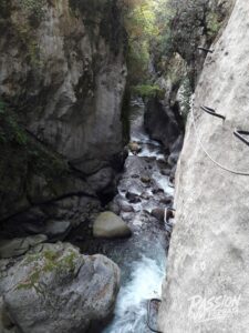 Narrow canyon with steep rocky walls, a fast-flowing stream at the bottom, and a metal ladder railing climbing along the right rock face