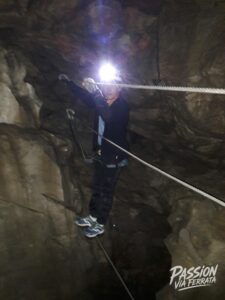 Person wearing a headlamp and safety gear traverses a rocky cave wall with ropes overhead.